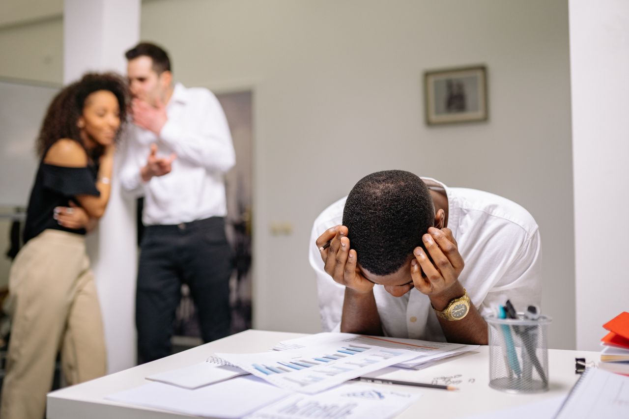 The Art of Drawing Readers In: Your attractive post title goes here Despairing worker at office desk as colleagues gossip in background illustrating workplace bullying.