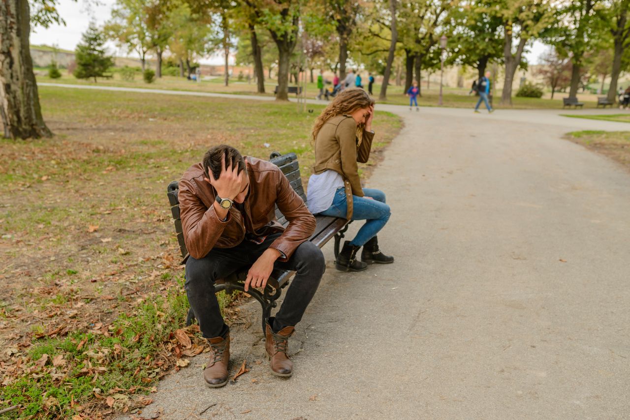Distraught couple sitting apart on a park bench, facing away from each other, in a public park setting.